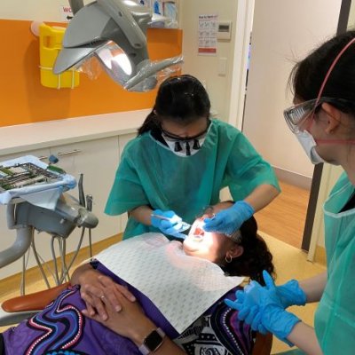 A patient lies on a reclining chair in a dental clinic as two student dentists in scrubs treat her.